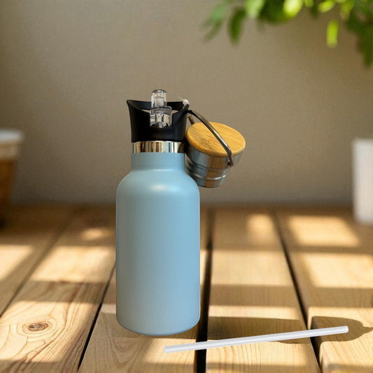 Hellblaue Nomafu Edelstahl Trinkflasche 350 ml mit Sportaufsatz, Strohhalm und Holzdeckel auf einem Holztisch