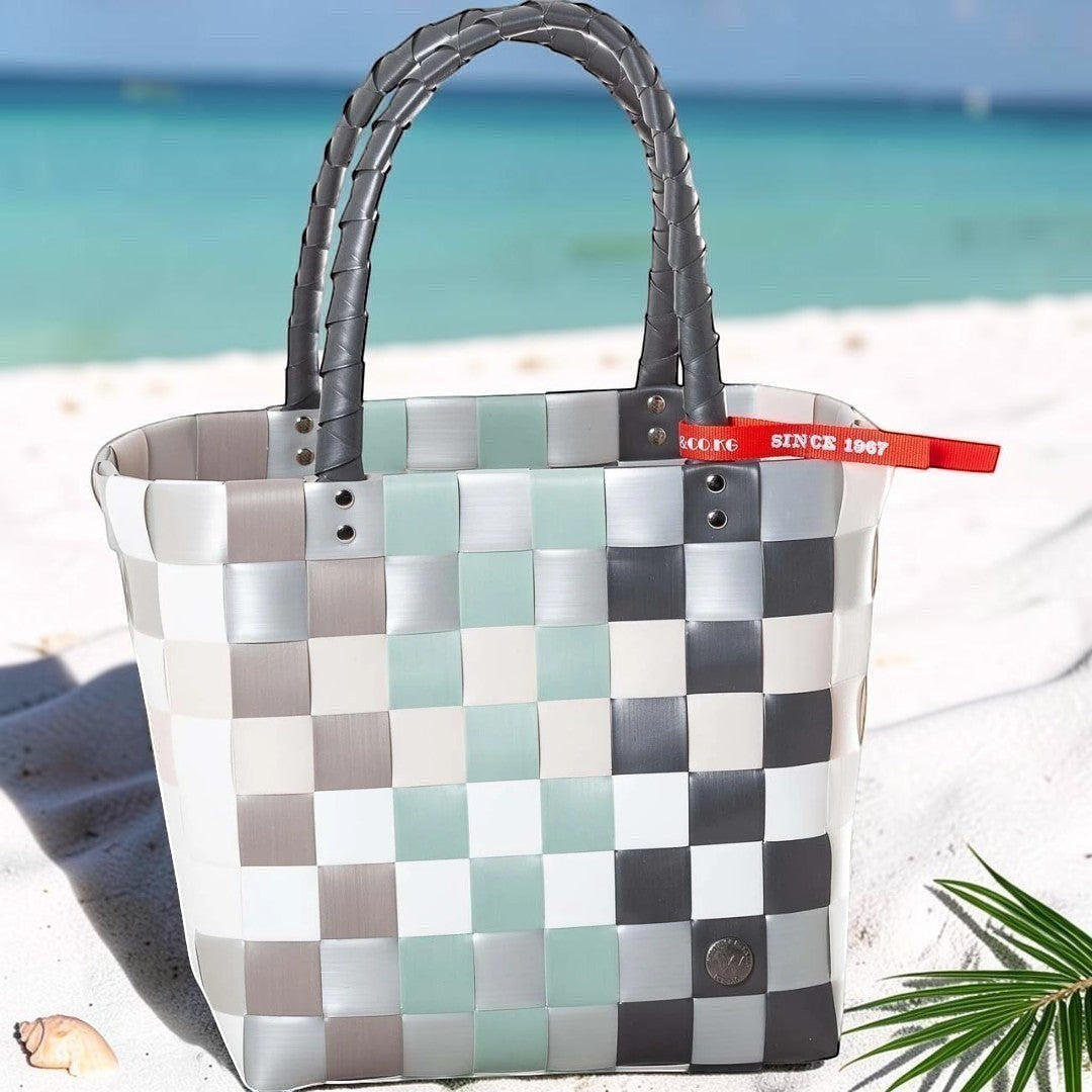 Nomafu geflochtene Tragetasche in Grau, Weiß und Mint mit rotem Detail am Strand, aus strapazierfähigem Kunststoff.