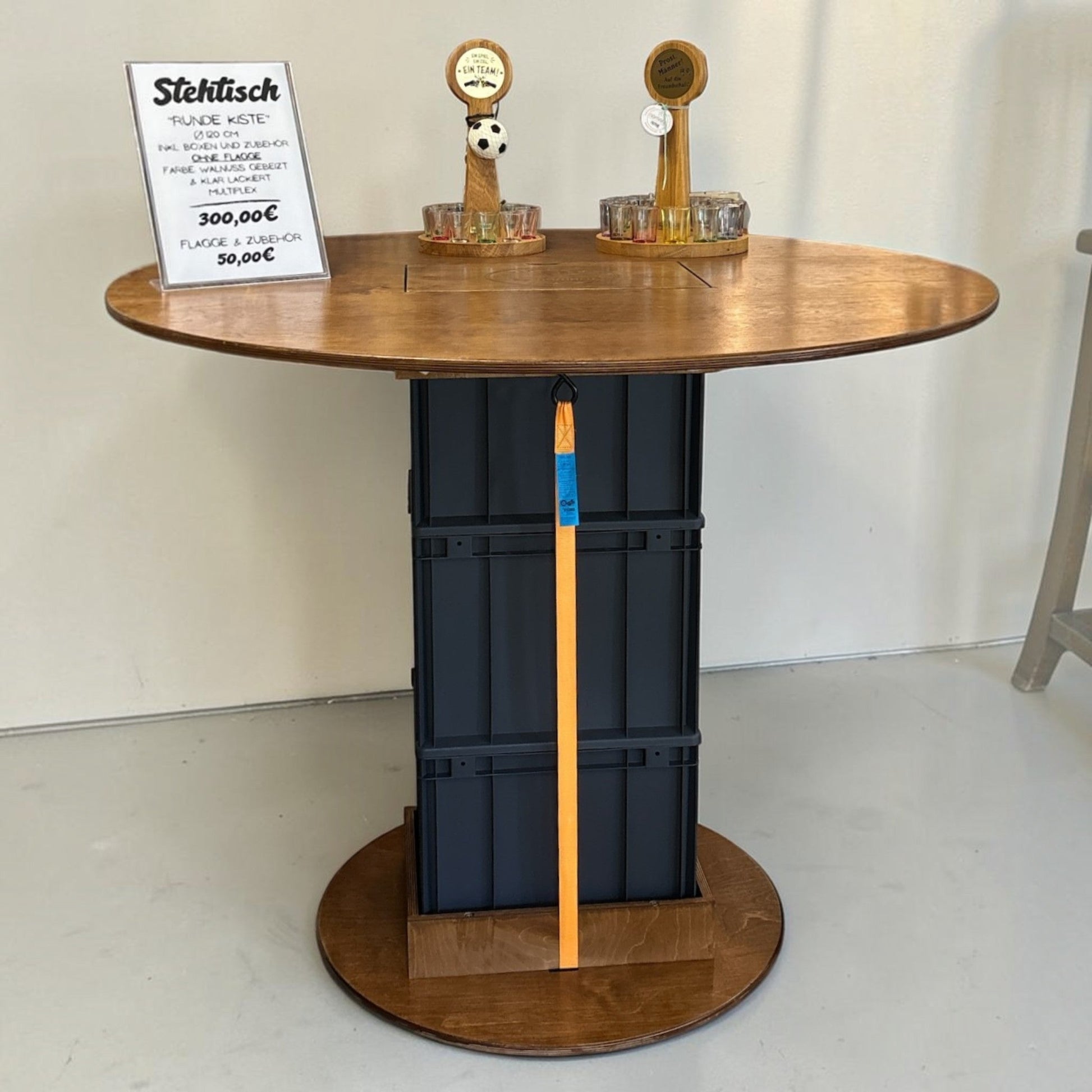 Nomafu Runde Kiste XXL round standing table with walnut-stained multiplex top, black beverage crates, and orange strap.
