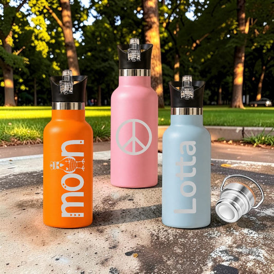 Three Nomafu stainless steel water bottles in orange, pink, and light blue with custom engravings in a park setting.